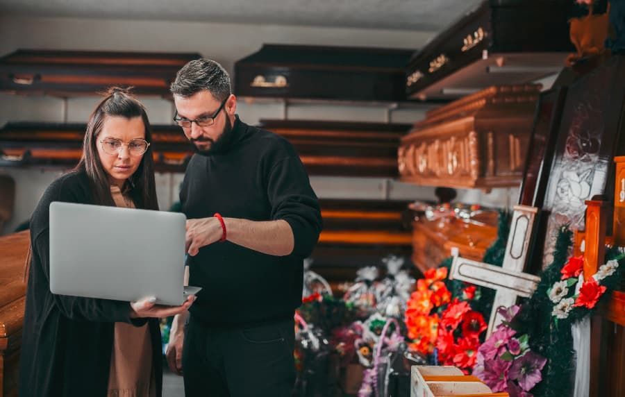 Funeral home workers coordinating on laptop with flowers and caskets behind them