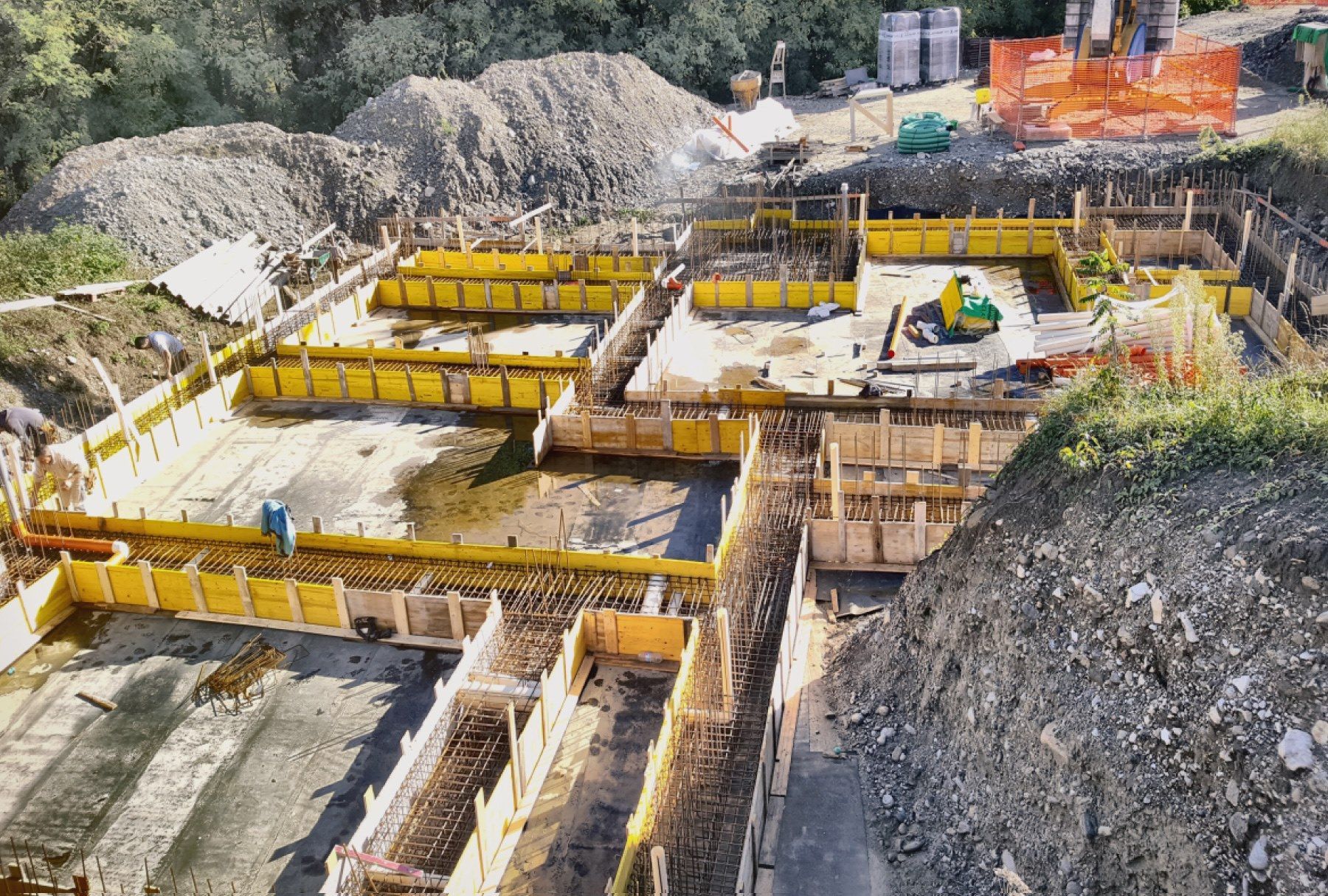 Cantiere edile con fondamenta in cemento, casseforme gialle e trincee scavate nel terreno.