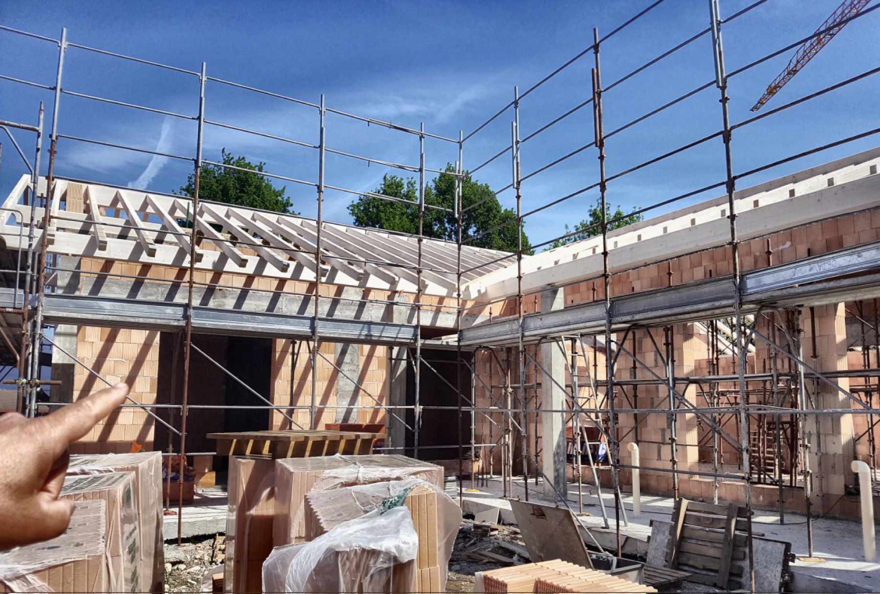 Cantiere edile con struttura in legno, impalcature e una mano che indica l'edificio sotto un cielo azzurro.
