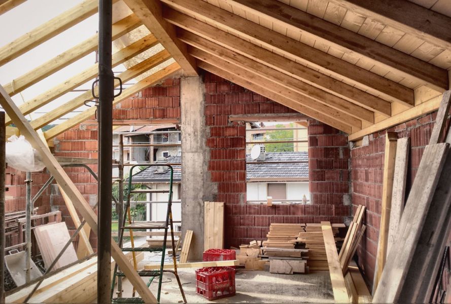 Struttura del sottotetto con travi in legno a vista, muri in mattoni, scala e materiali accatastati sul pavimento.