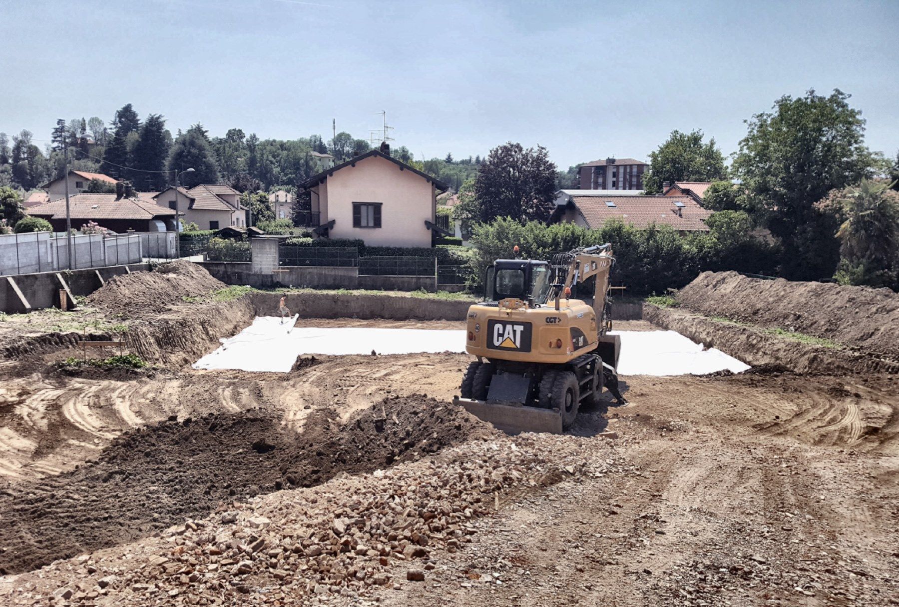 Un escavatore CAT giallo sposta terra in un cantiere edile accanto alle case, sotto un cielo sereno.