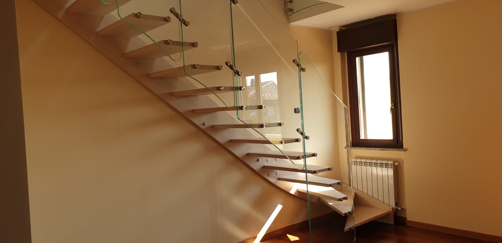 Scala moderna con ringhiera in vetro in una stanza beige, accanto a una finestra e a un termosifone.