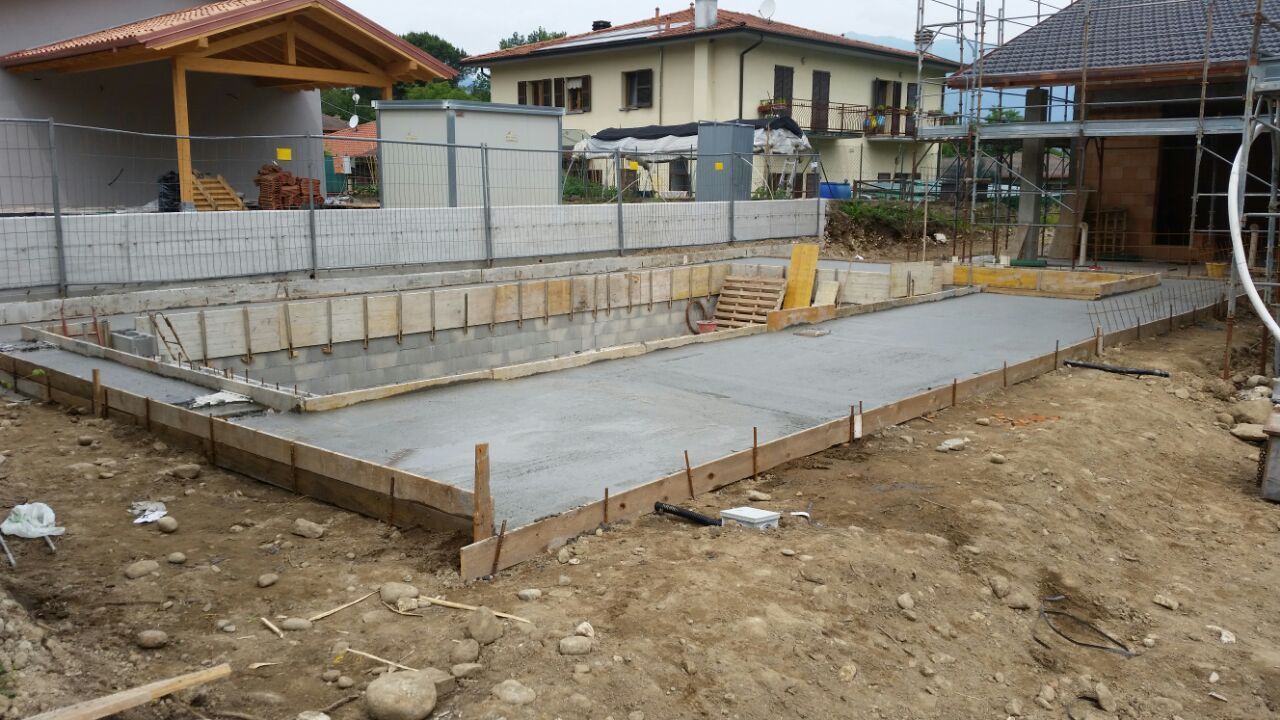 Fondamenta in cemento in costruzione su un terreno sterrato, con armature in ferro e case sullo sfondo.