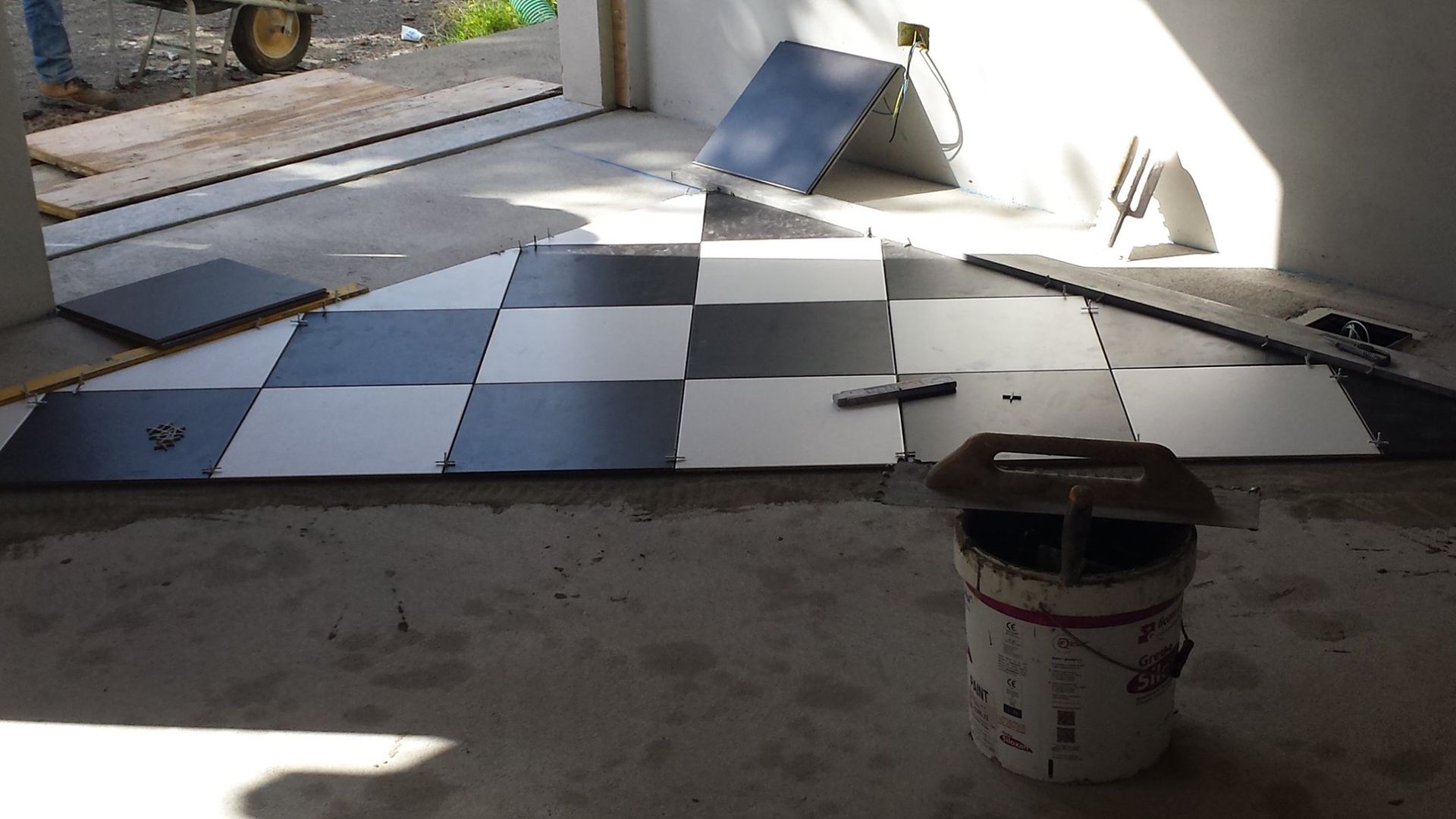 In una stanza soleggiata, si sta installando un pavimento a scacchiera bianco e nero, con un secchio di vernice in primo piano.