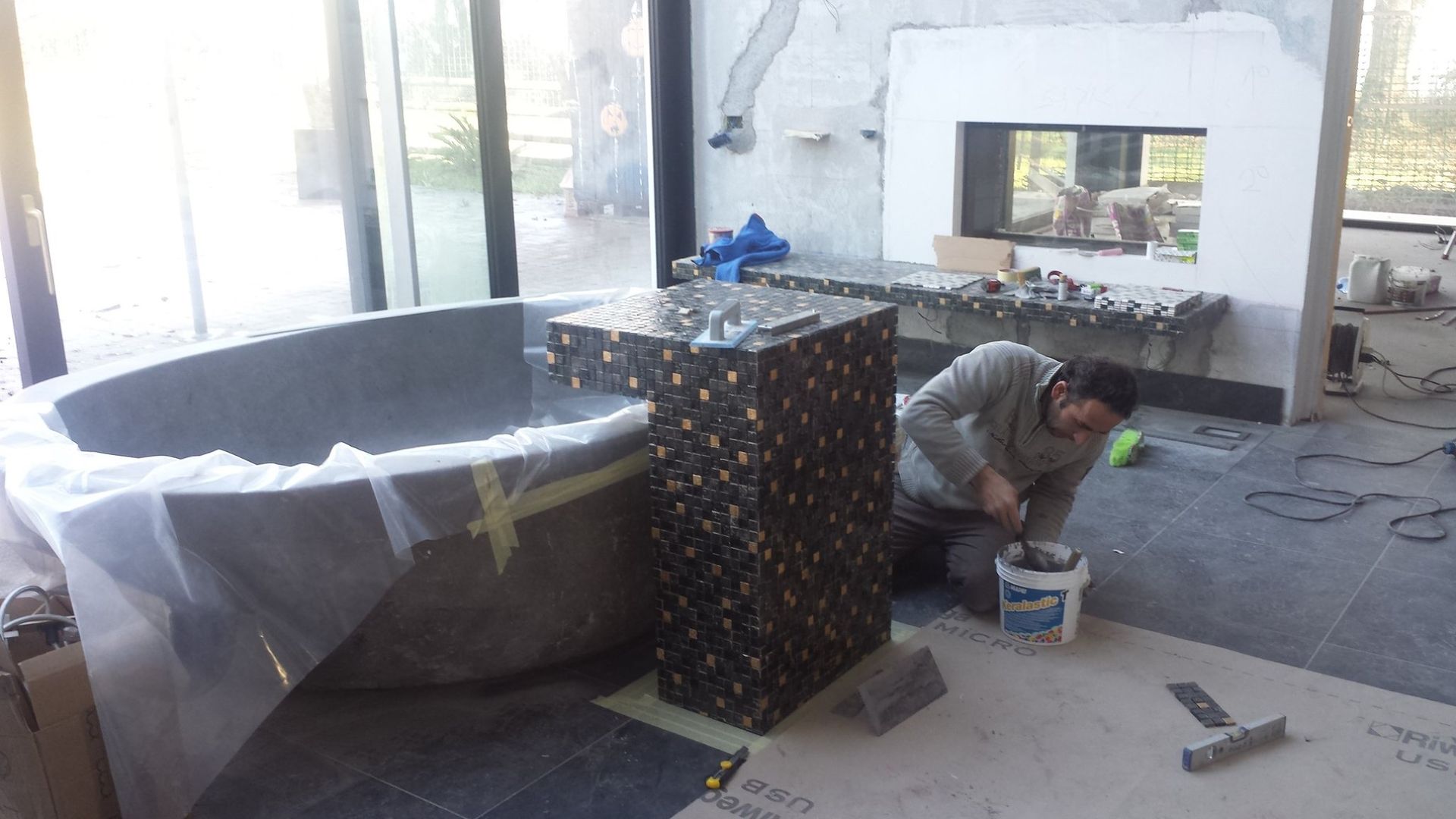 Operaio inginocchiato su un pavimento piastrellato, intento a installare un bancone a mosaico in una stanza luminosa.