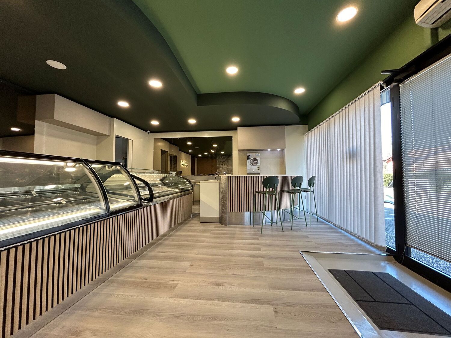 Interno moderno di una caffetteria con vetrine, bancone a righe, finestre alte e una piccola area con posti a sedere.