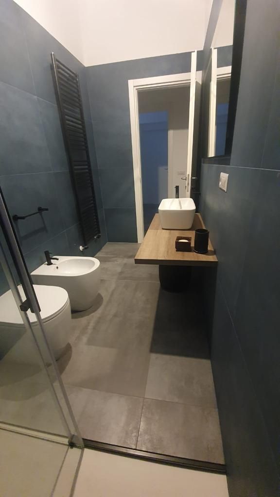 Bagno moderno con doccia in vetro, WC sospeso, piastrelle grigie e mobiletto in legno con lavabo e specchio.