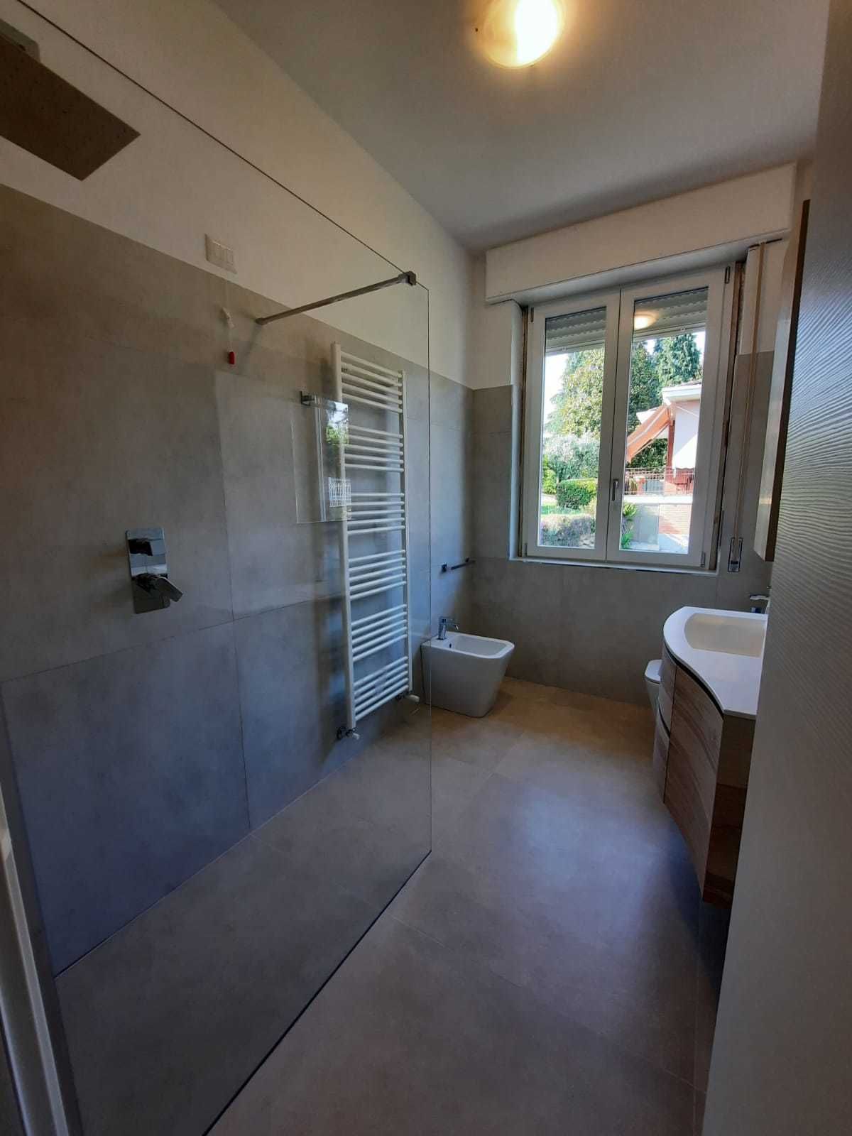 Bagno moderno con doccia in vetro, lavabo con mobiletto, WC e finestra con vista sull'esterno.