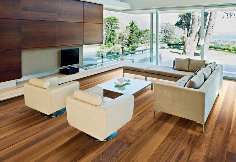 Soggiorno moderno con divano angolare beige, sedie bianche, pavimento in legno e ampie finestre che si affacciano sul verde.