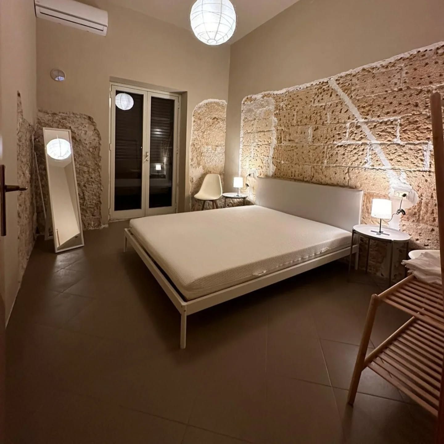 Accogliente camera da letto beige con letto, lampade, finestra, specchio e sedia in vimini sotto una decorazione murale intrecciata.
