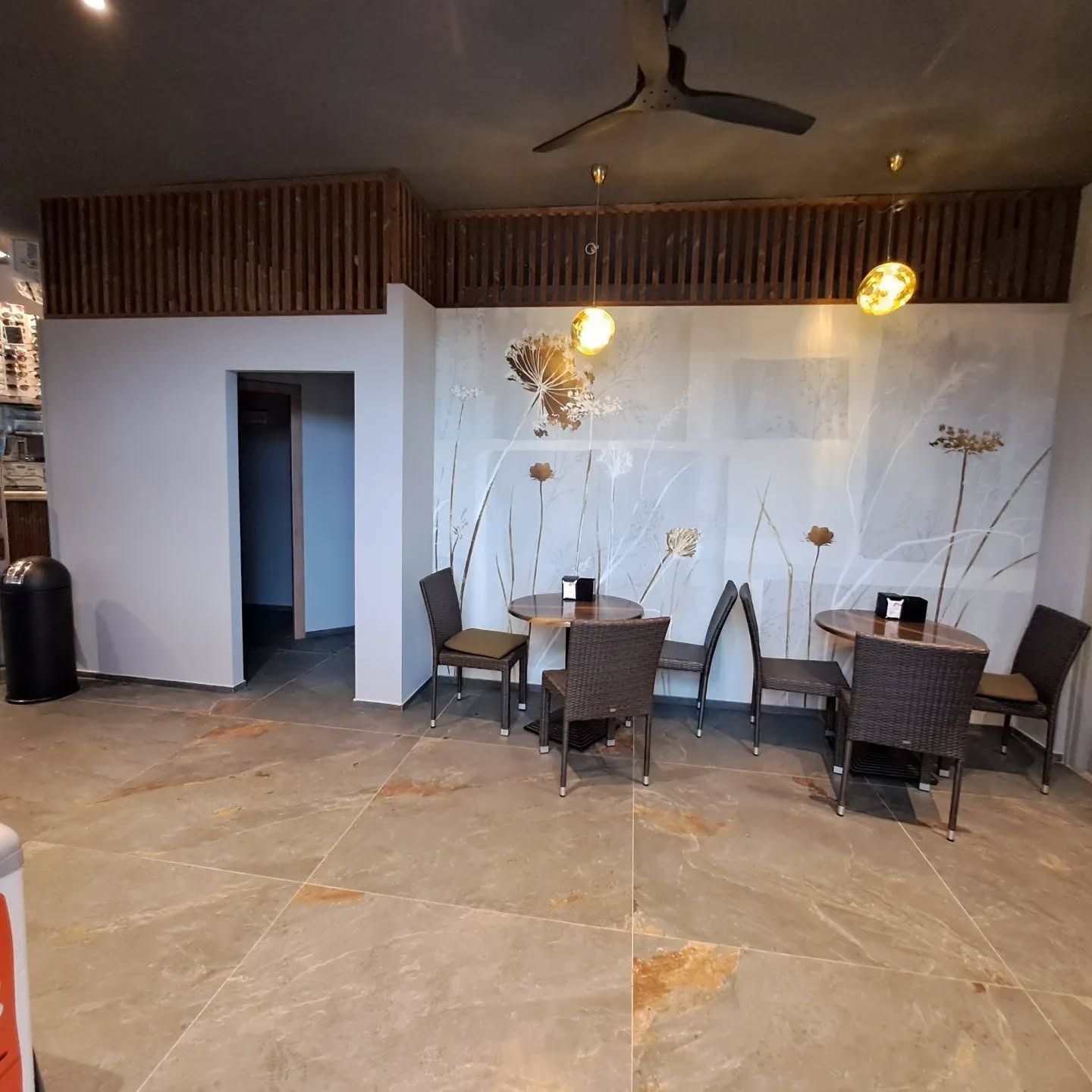 Interno moderno di un caffè con sedie in vimini, tavolini e una parete bianca con rami decorativi.