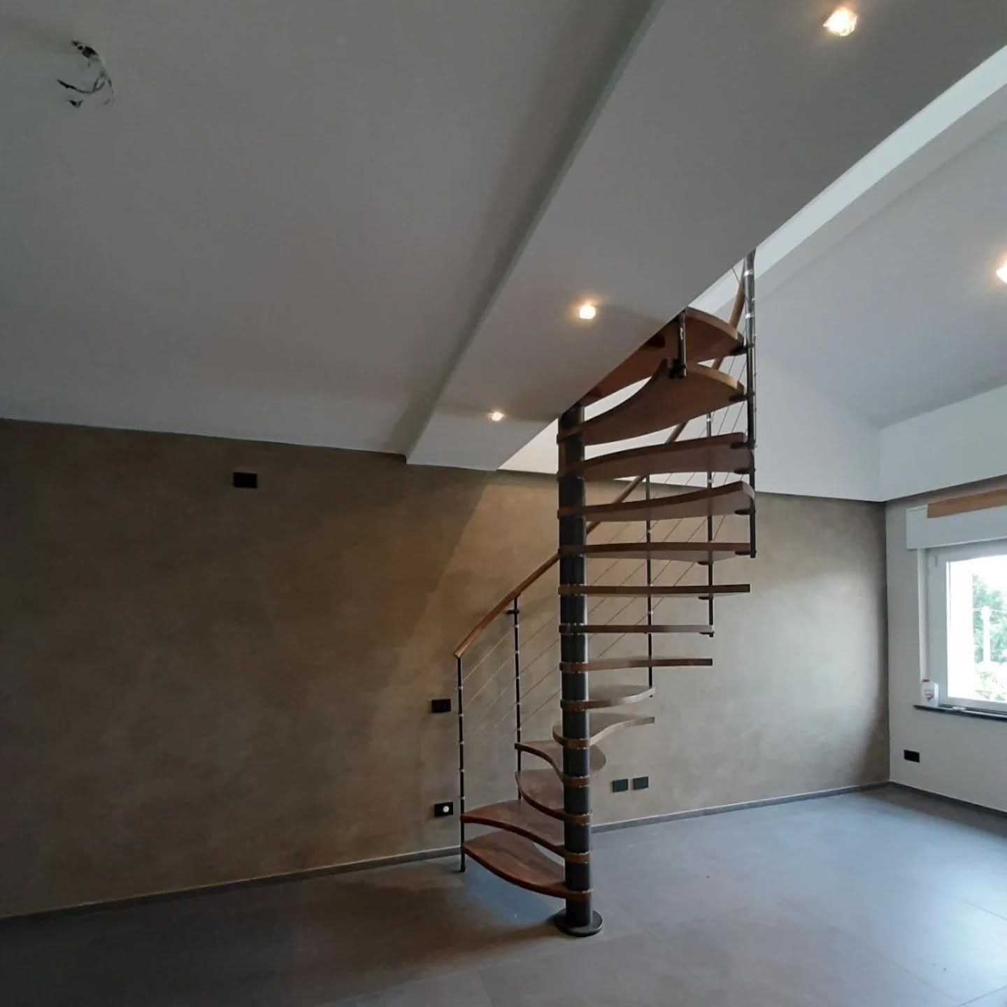 Camera moderna con scala a chiocciola, pareti beige e faretti a incasso sul soffitto.