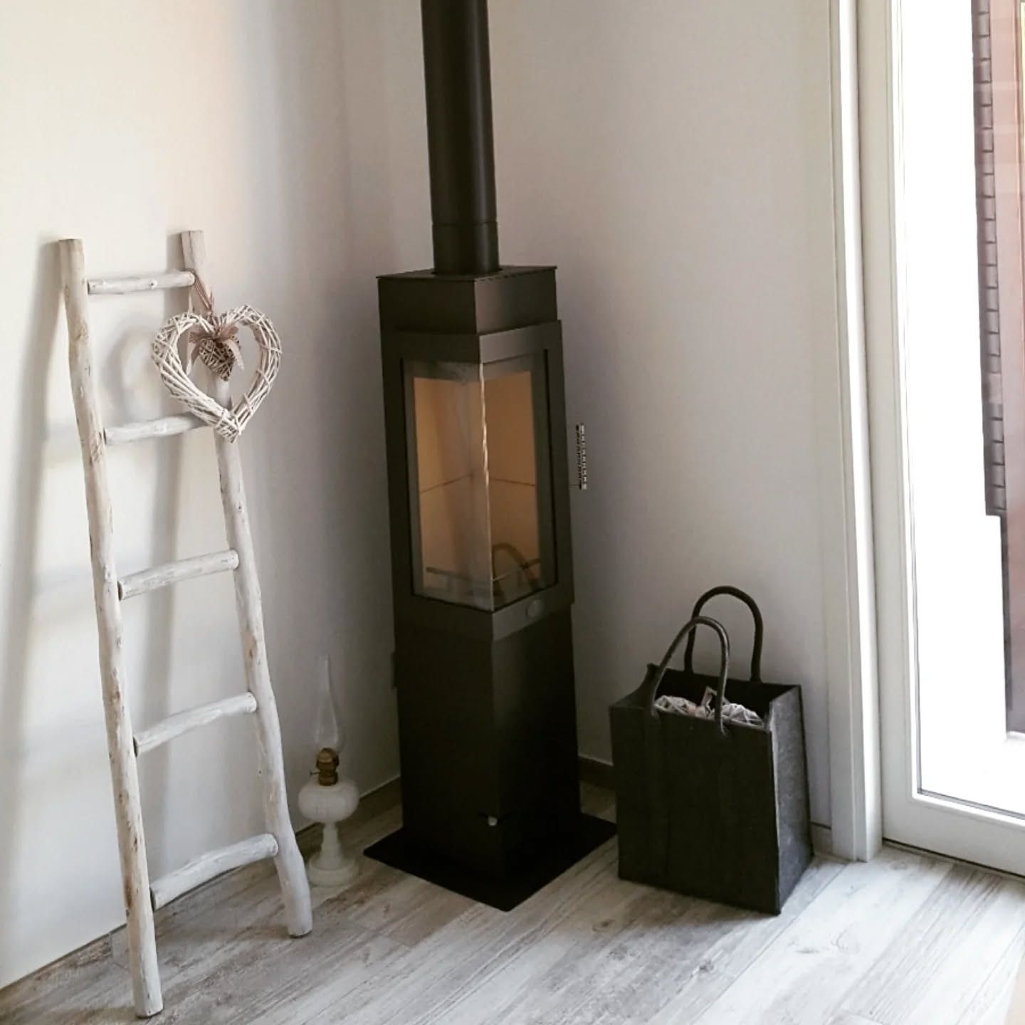 Angolo di una stanza moderna con stufa a legna nera, mensola a scala bianca e borsa verde accanto a una porta a vetri.