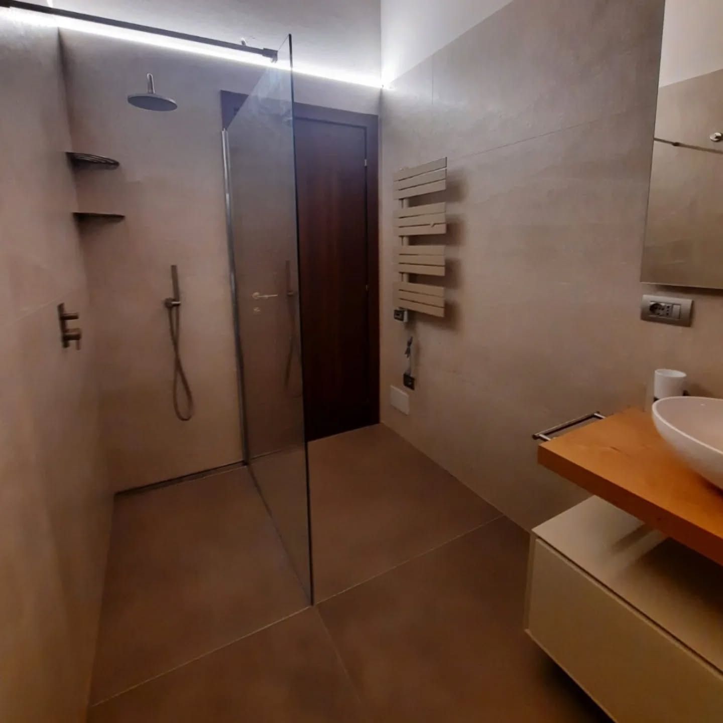 Bagno moderno con doccia in vetro, pareti rivestite con piastrelle beige, mobile lavabo in legno e termoarredo portasciugamani.
