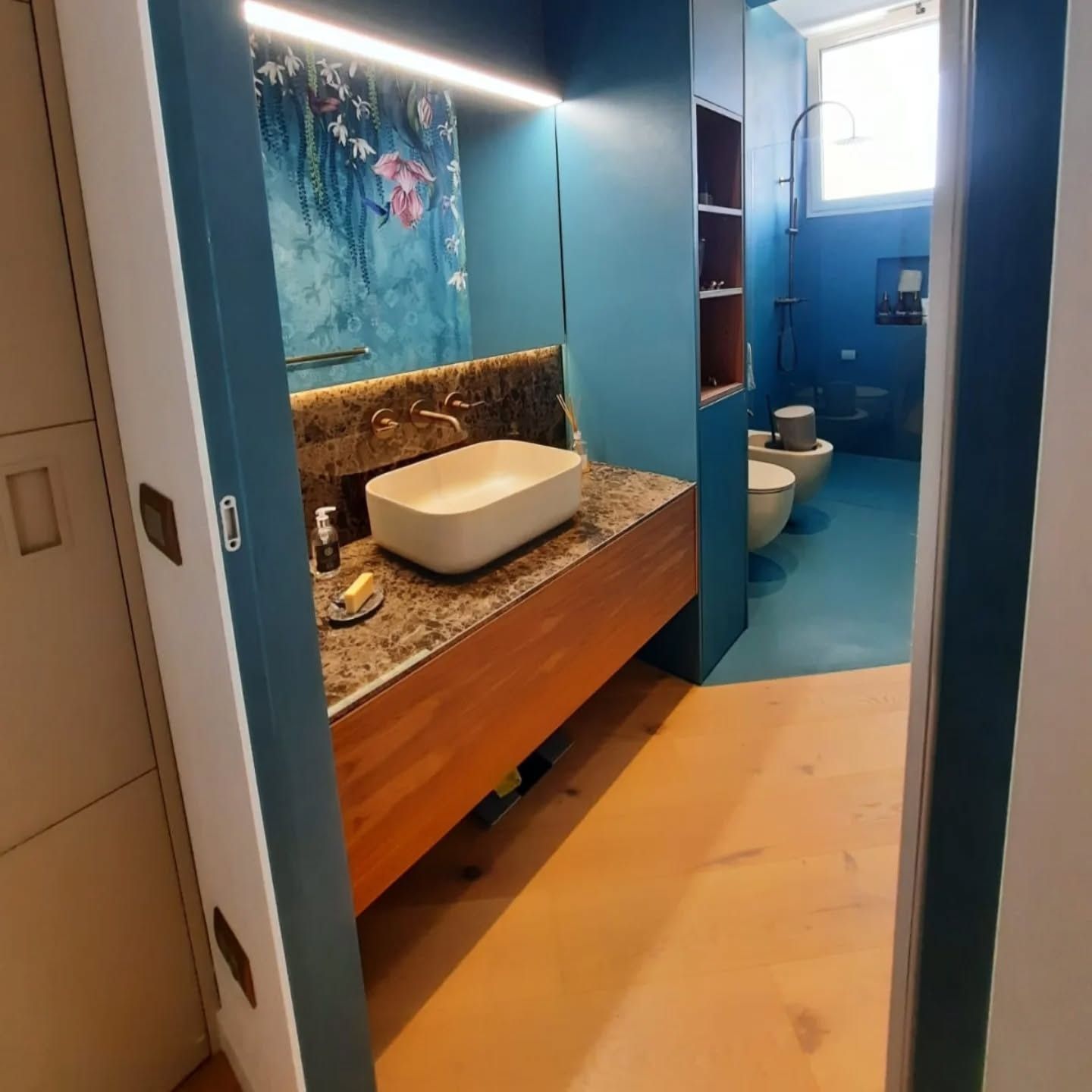 Bagno moderno con pareti blu, mobiletto sospeso in legno, lavabo bianco e WC visibile attraverso la porta aperta.