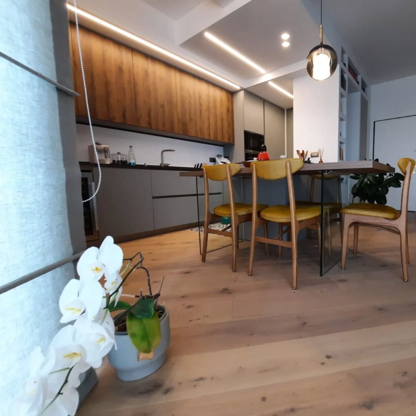 Cucina moderna a vista con zona pranzo, tavolo in legno, sedie gialle e orchidea in primo piano.