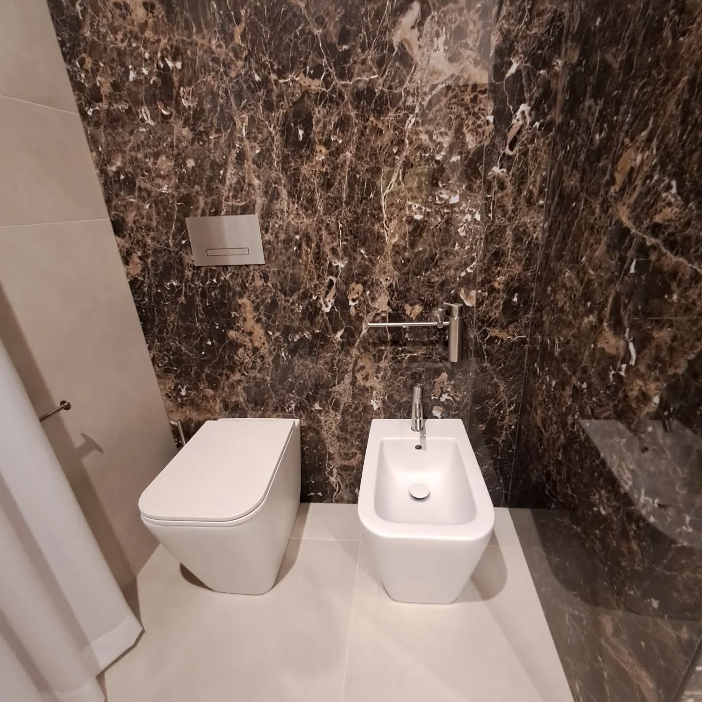 Bagno moderno con pareti in marmo scuro, WC sospeso, bidet e porta doccia in vetro.