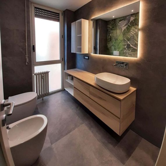 Bagno moderno con mobile lavabo sospeso in legno, lavabo a ciotola, specchio, bidet, WC e finestra satinata.
