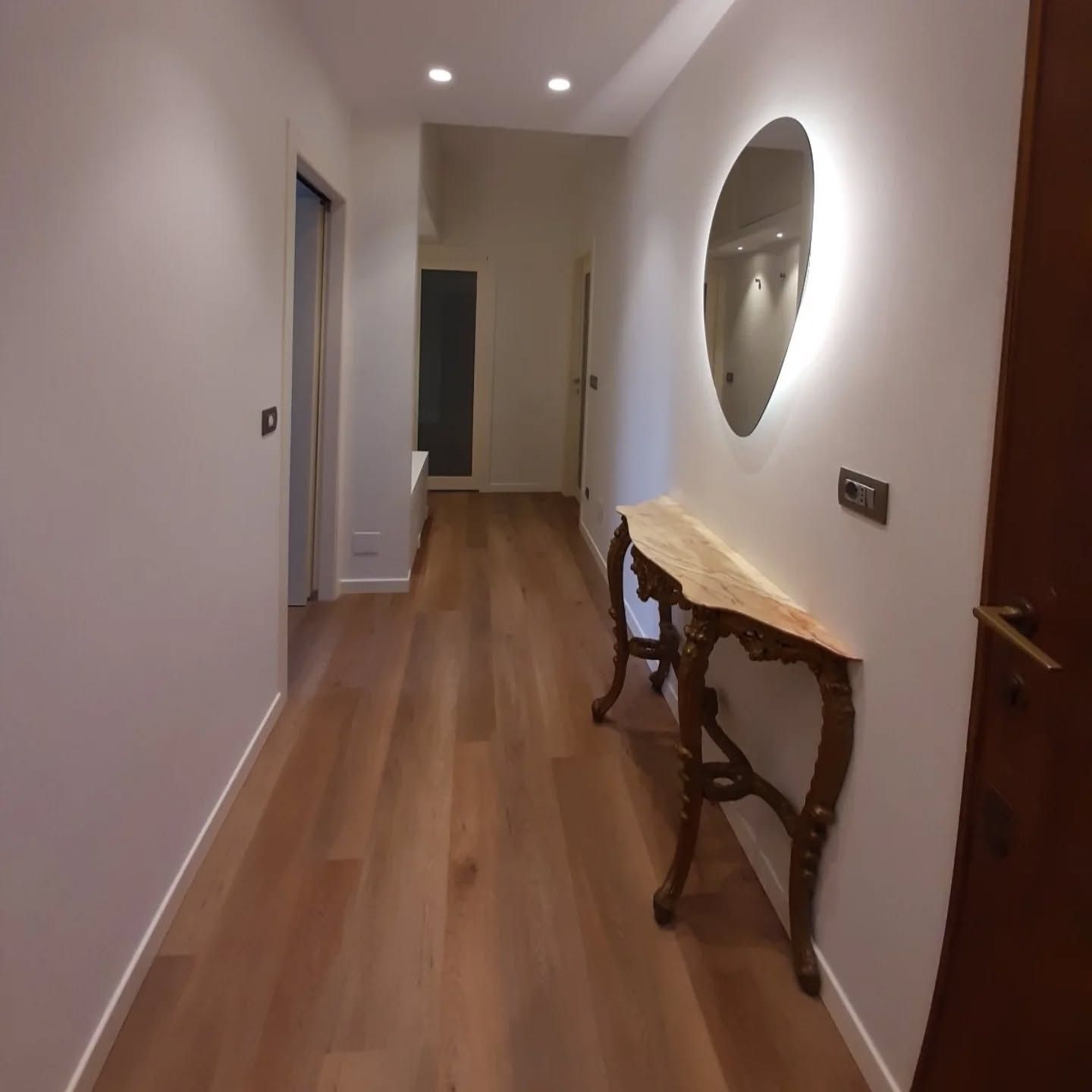 Luminoso corridoio con pavimento in legno, pareti bianche, consolle e specchio ovale.