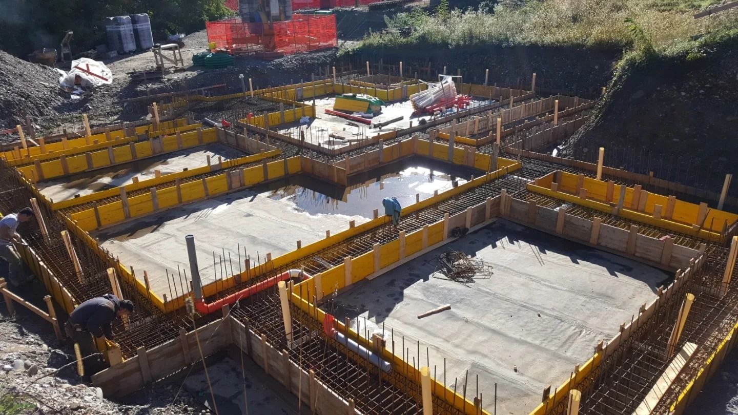 Cantiere edile con lastre di fondazione in cemento, casseforme gialle e ponteggi attorno a una fossa di costruzione.
