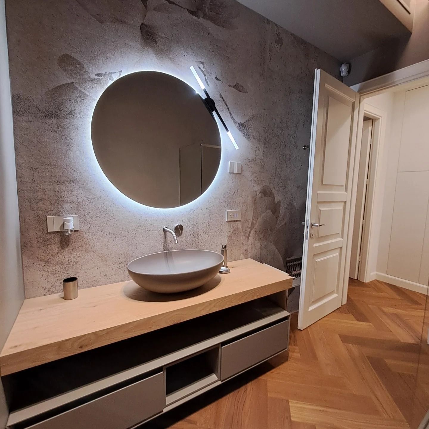 Bagno moderno con specchio rotondo retroilluminato, lavabo a ciotola, mobiletto in legno e pavimento in legno a spina di pesce.