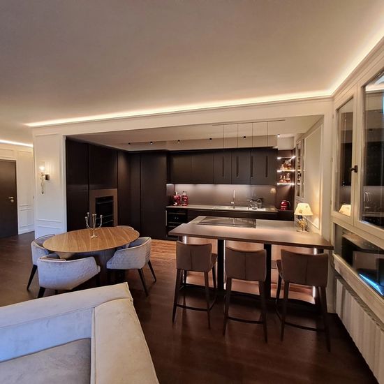 Cucina e zona pranzo moderne con mobili scuri, sgabelli a isola e un tavolo rotondo in legno in un ambiente open space illuminato da una luce calda.