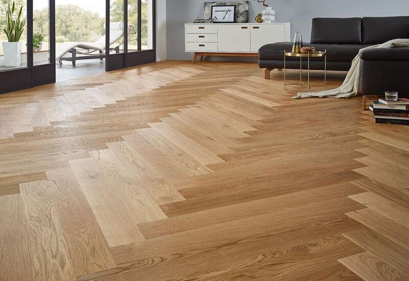 Un luminoso pavimento in legno a spina di pesce in un soggiorno moderno con divano grigio e ampie finestre.
