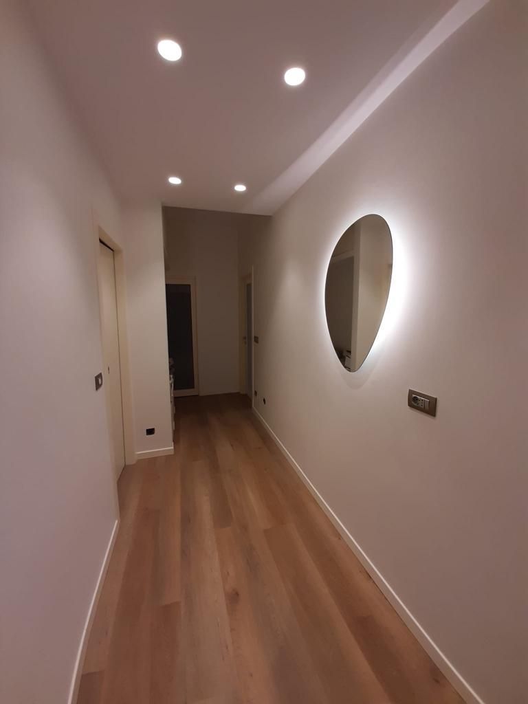 Stretto corridoio con pavimento in legno, faretti a incasso sul soffitto e specchio rotondo a parete.