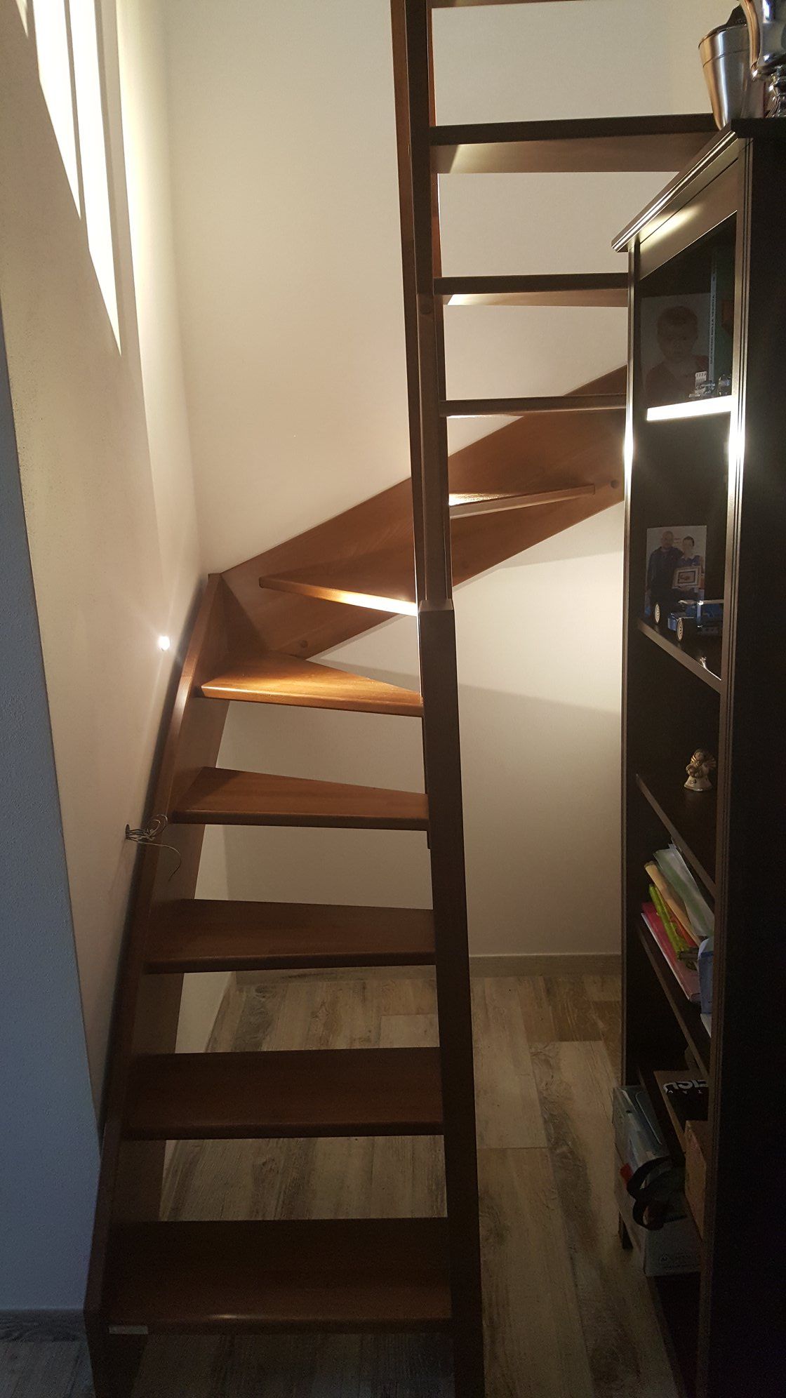 Stretta scala in legno con una ripida curva accanto a una libreria in un interno poco illuminato.