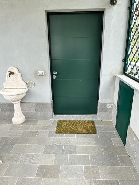 Soggiorno minimalista con porta beige, orologio a parete, piante, sedia e divano accanto a una finestra con cornice nera.
