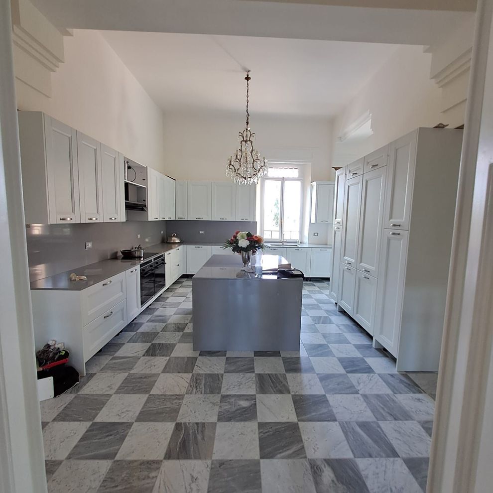 Cucina luminosa e bianca con pavimento a scacchi grigi, isola centrale, lampadario e pensili alti.
