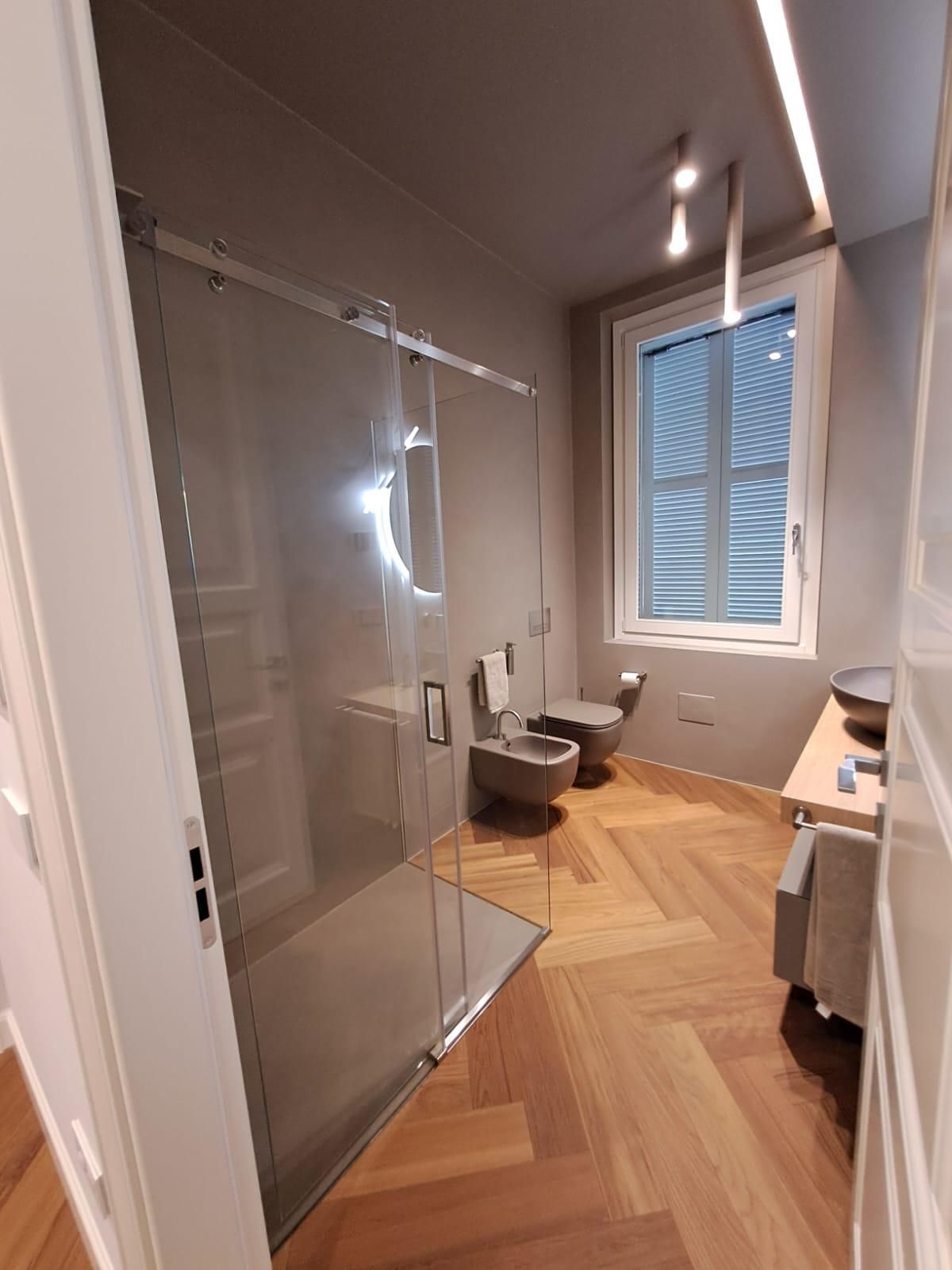 Bagno moderno con doccia in vetro, vasca freestanding, pavimento in legno e ampia finestra.