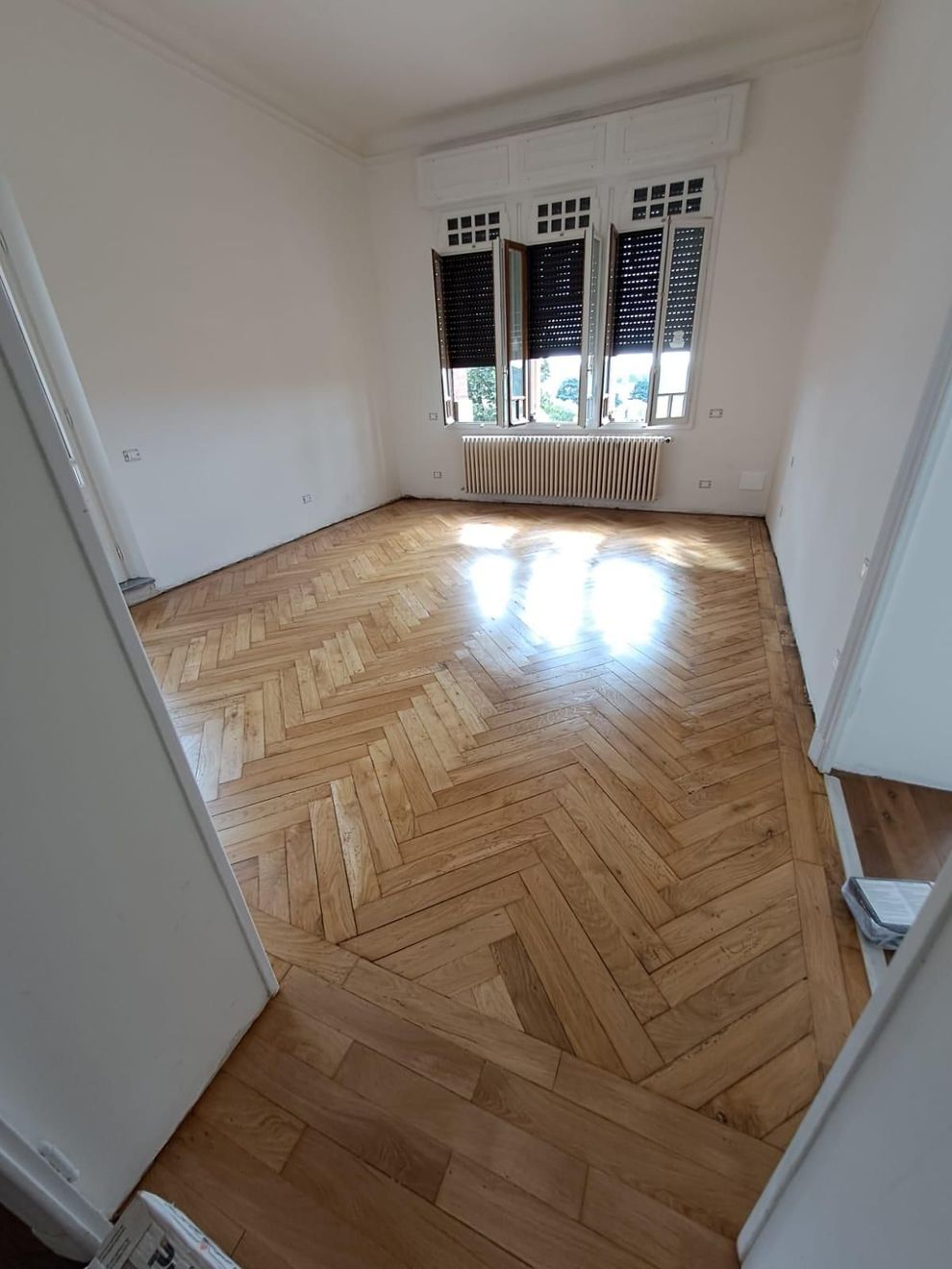 Stanza bianca vuota con pavimento in legno a spina di pesce, grande finestra e termosifone sotto la finestra.