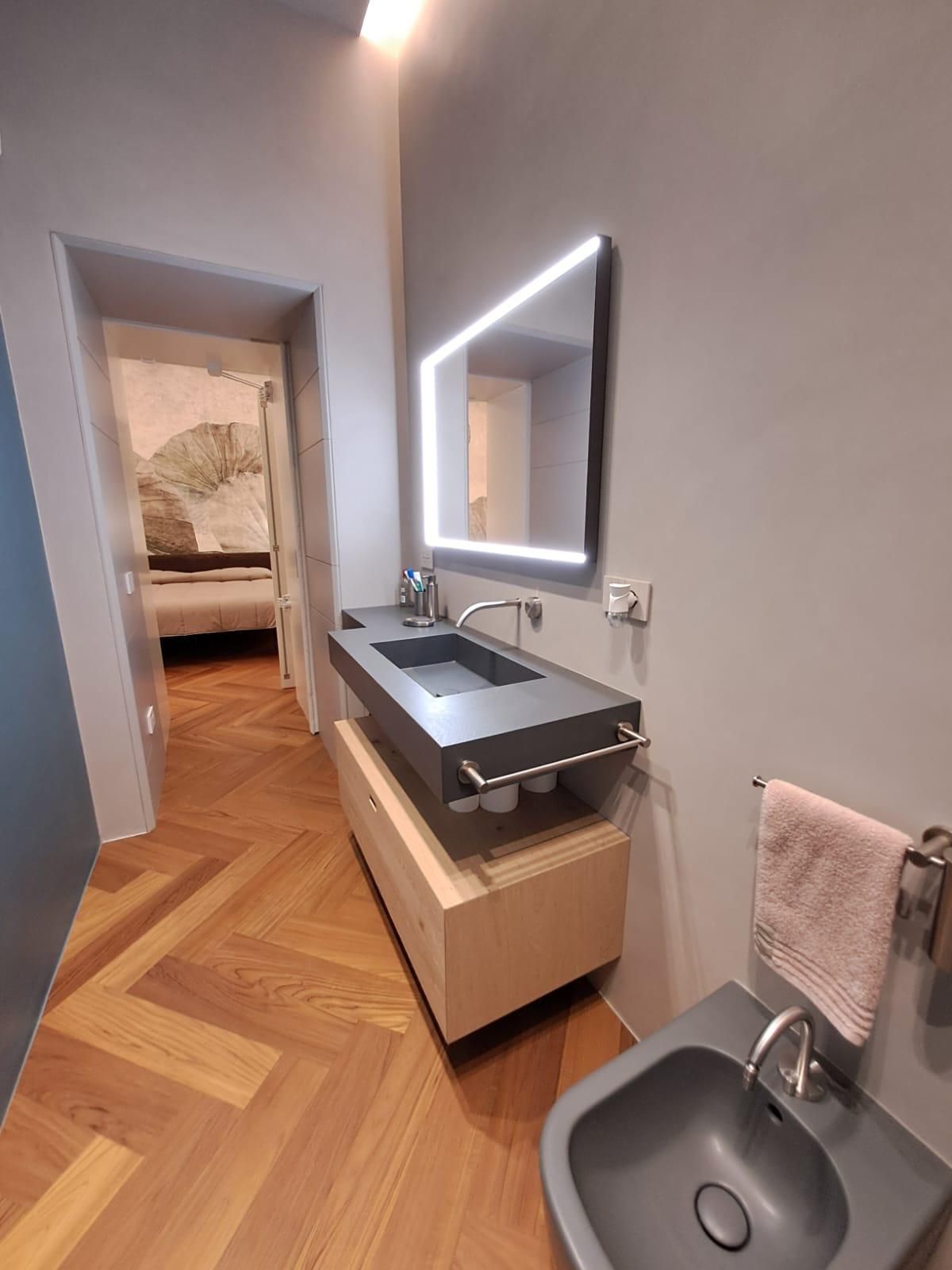 Bagno moderno con mobile sospeso, specchio illuminato, lavabo e portasciugamani; la porta si apre su una camera da letto.