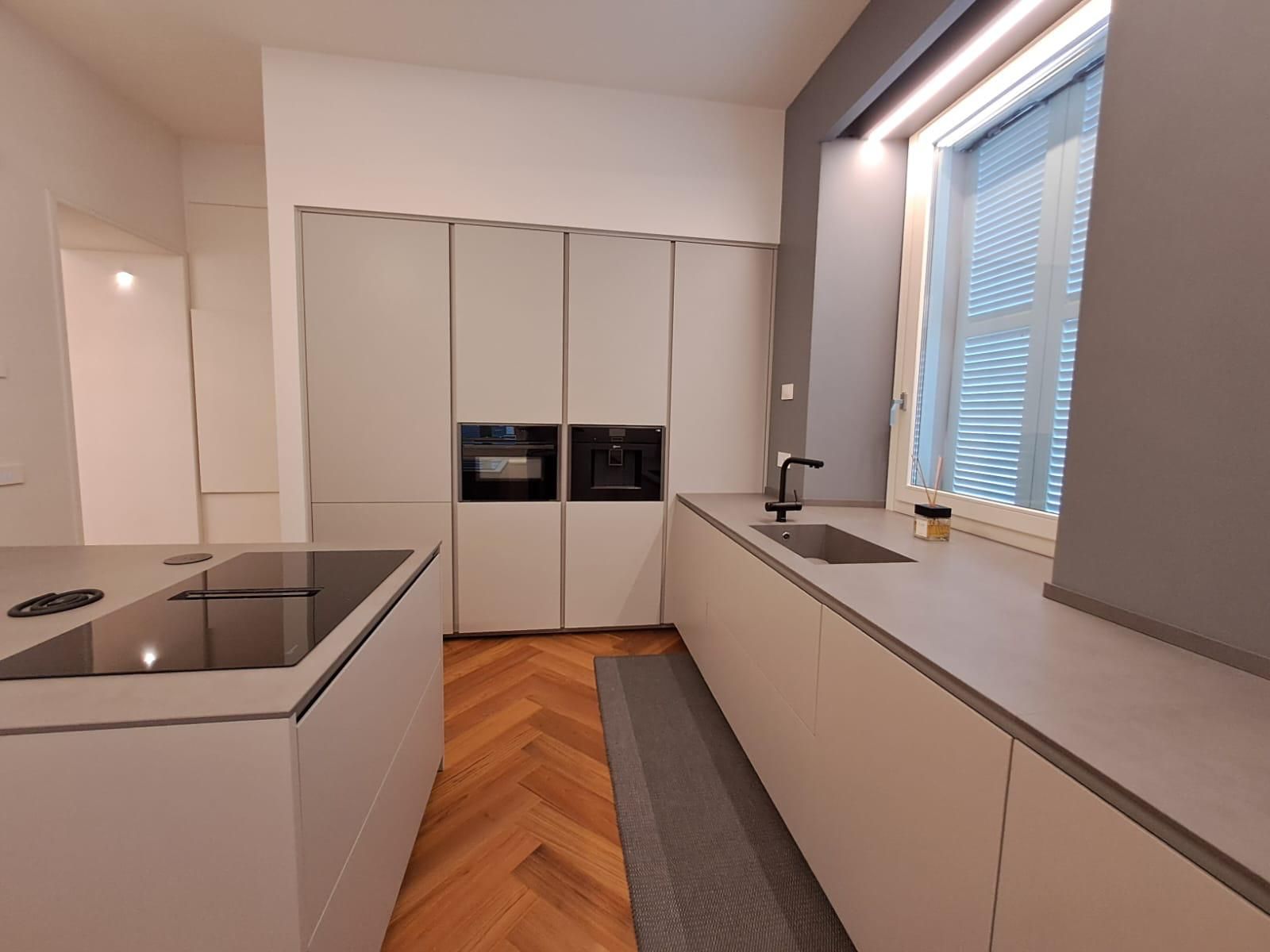 Cucina moderna e minimalista con mobili bianchi, isola con piano cottura, lavello vicino alla finestra e pavimento in legno a spina di pesce.