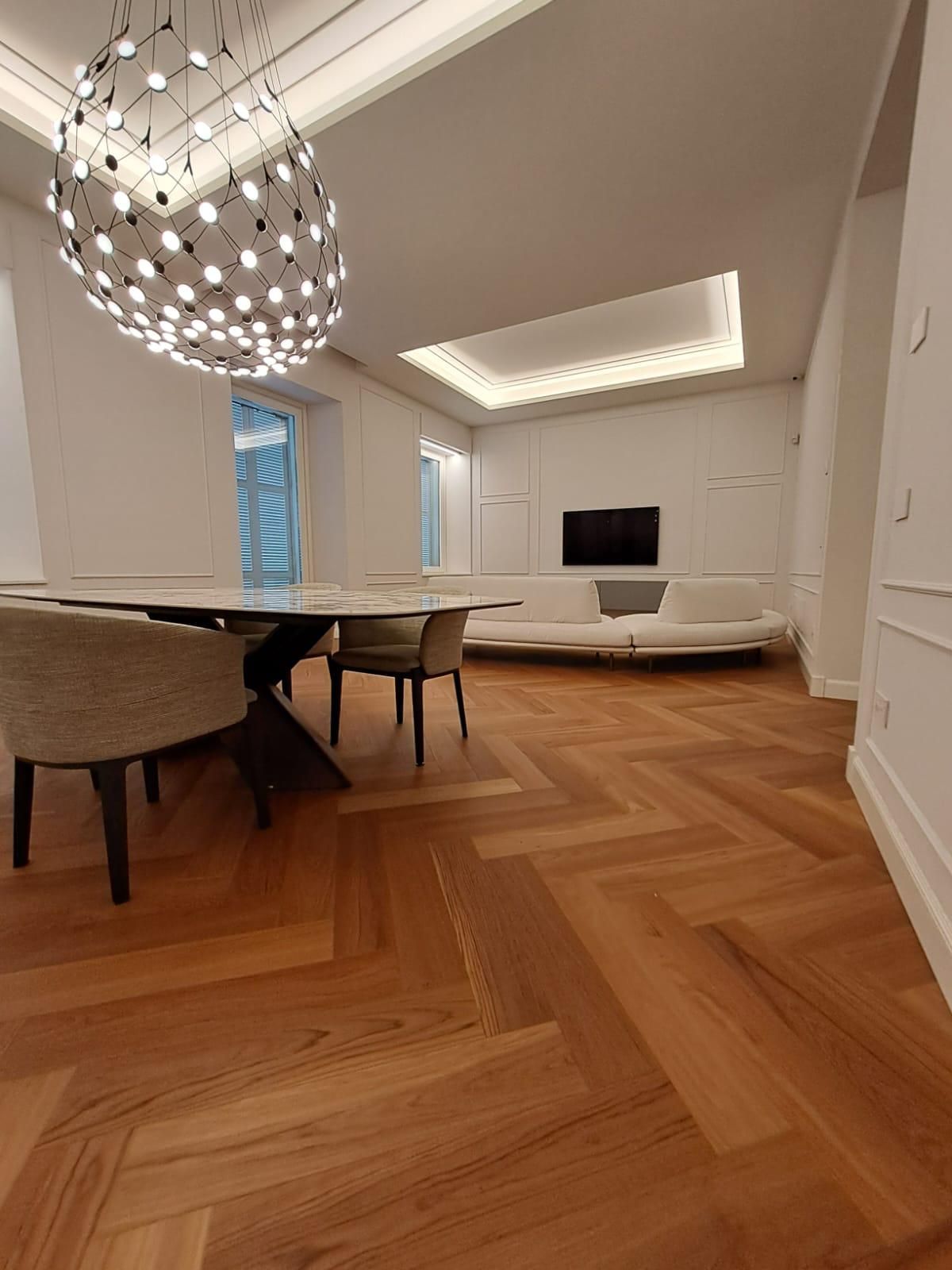 Soggiorno e sala da pranzo moderni con pavimento in legno a spina di pesce, lampadario, sedute bianche e TV a parete.