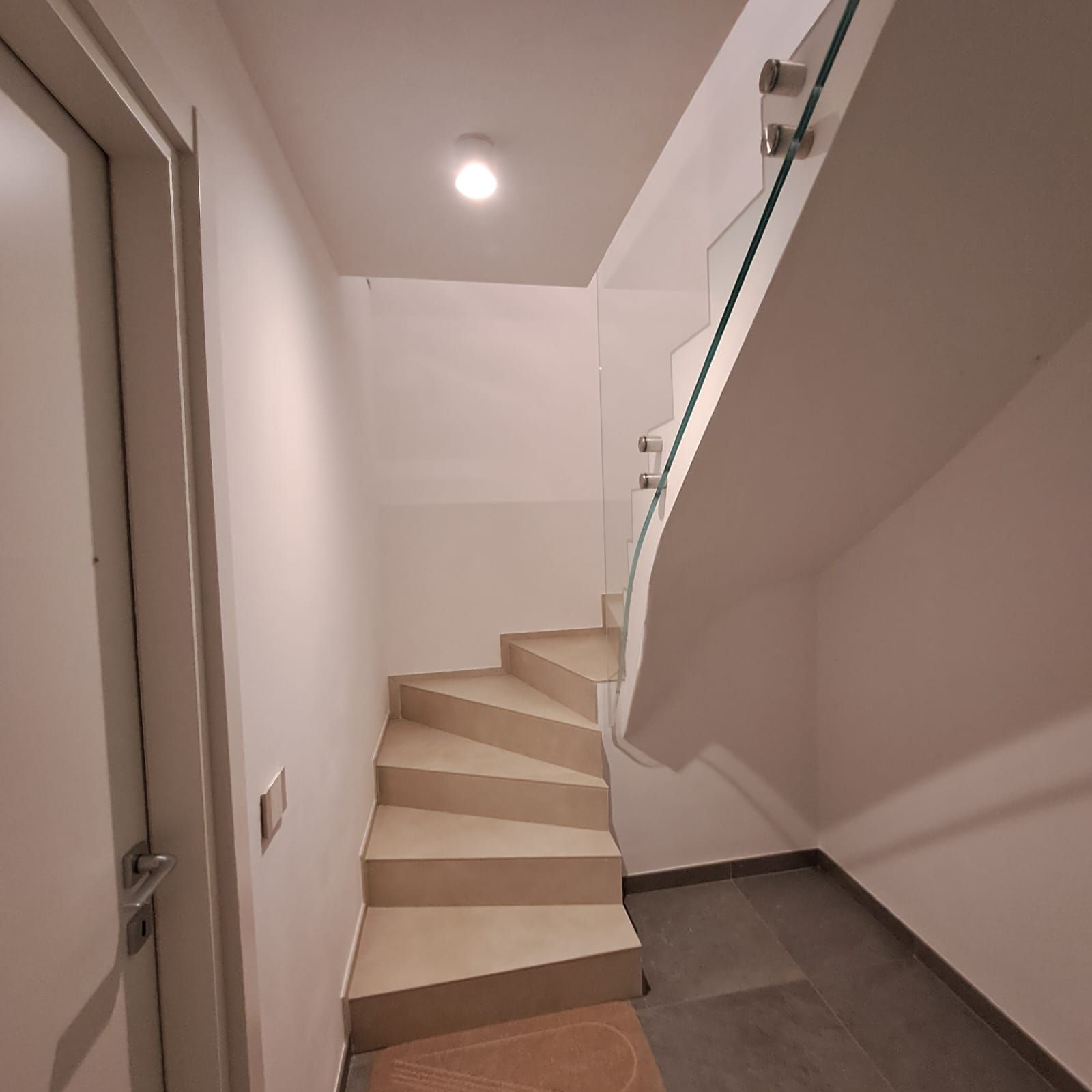 Scala interna stretta con gradini beige, pareti bianche e pianerottolo piastrellato in grigio sotto una lampada a soffitto.