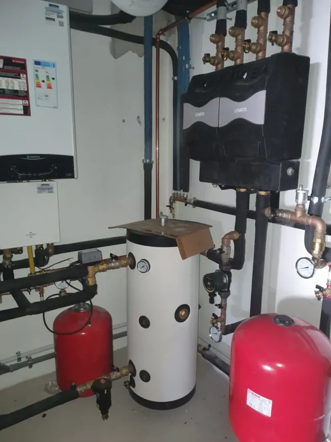 Locale caldaia con serbatoio bianco, vasi di espansione rossi, tubazioni e unità di controllo a parete.