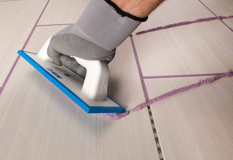 Mano guantata che utilizza uno strumento per la pulizia delle fughe su un pavimento piastrellato con fughe viola.