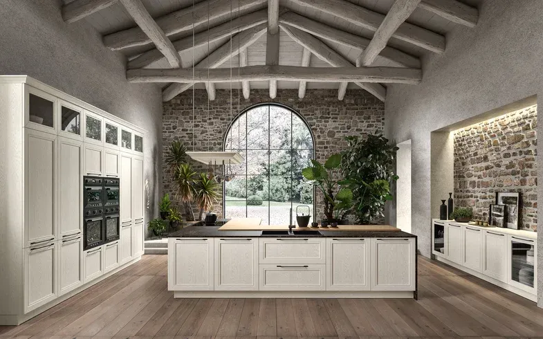 Cucina moderna e luminosa con mobili bianchi, isola centrale, finestra ad arco e travi in legno a vista.