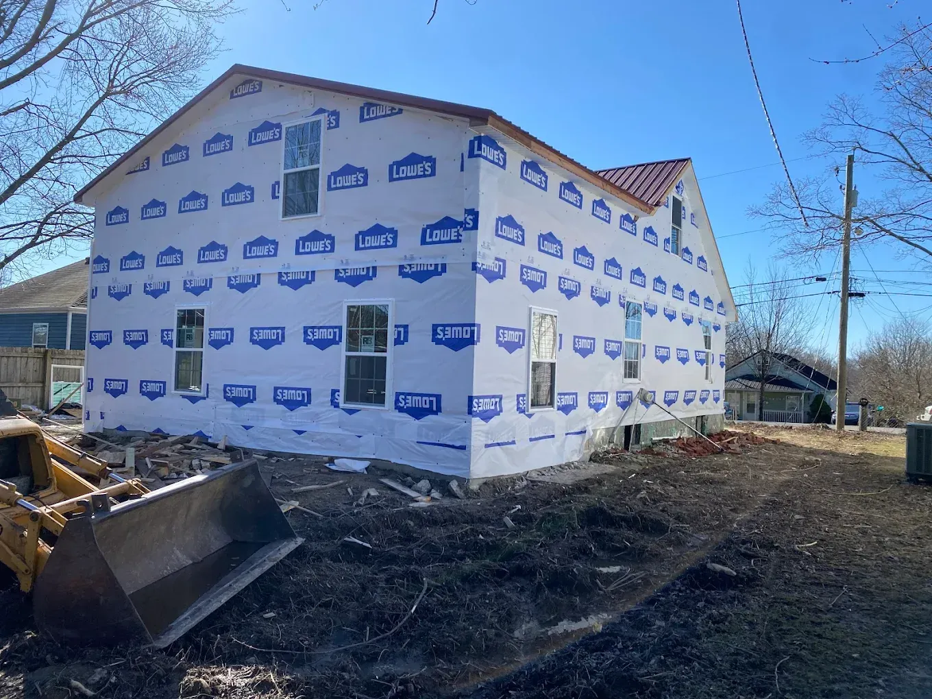A house under construction, covered in Lowe's house wrap. Dirt and an excavator in front.