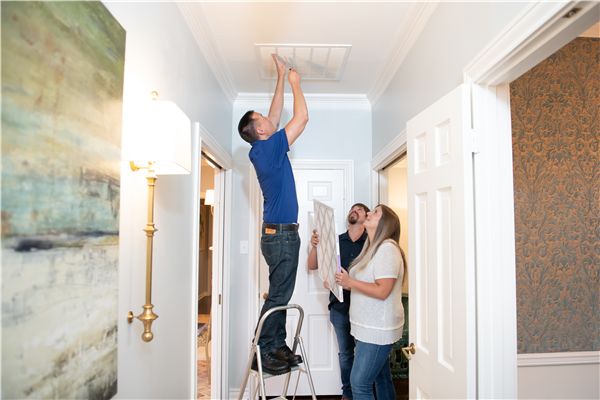 Two people on a ladder install a ceiling light in a bright hallway while another watches and assists