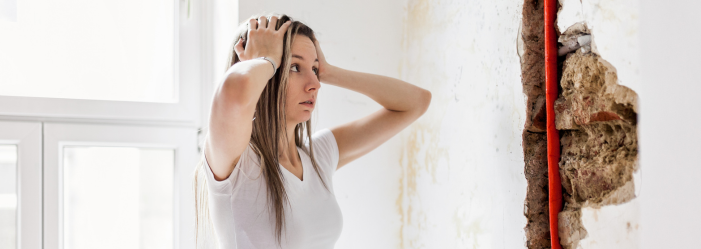 Person holding their head in a damaged wall area with exposed insulation and red wiring indoors