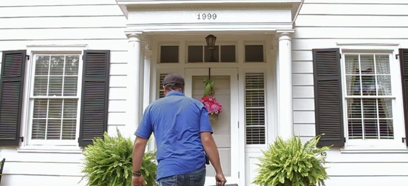 Man in blue shirt approaching a white house front door with potted plants and black shutters