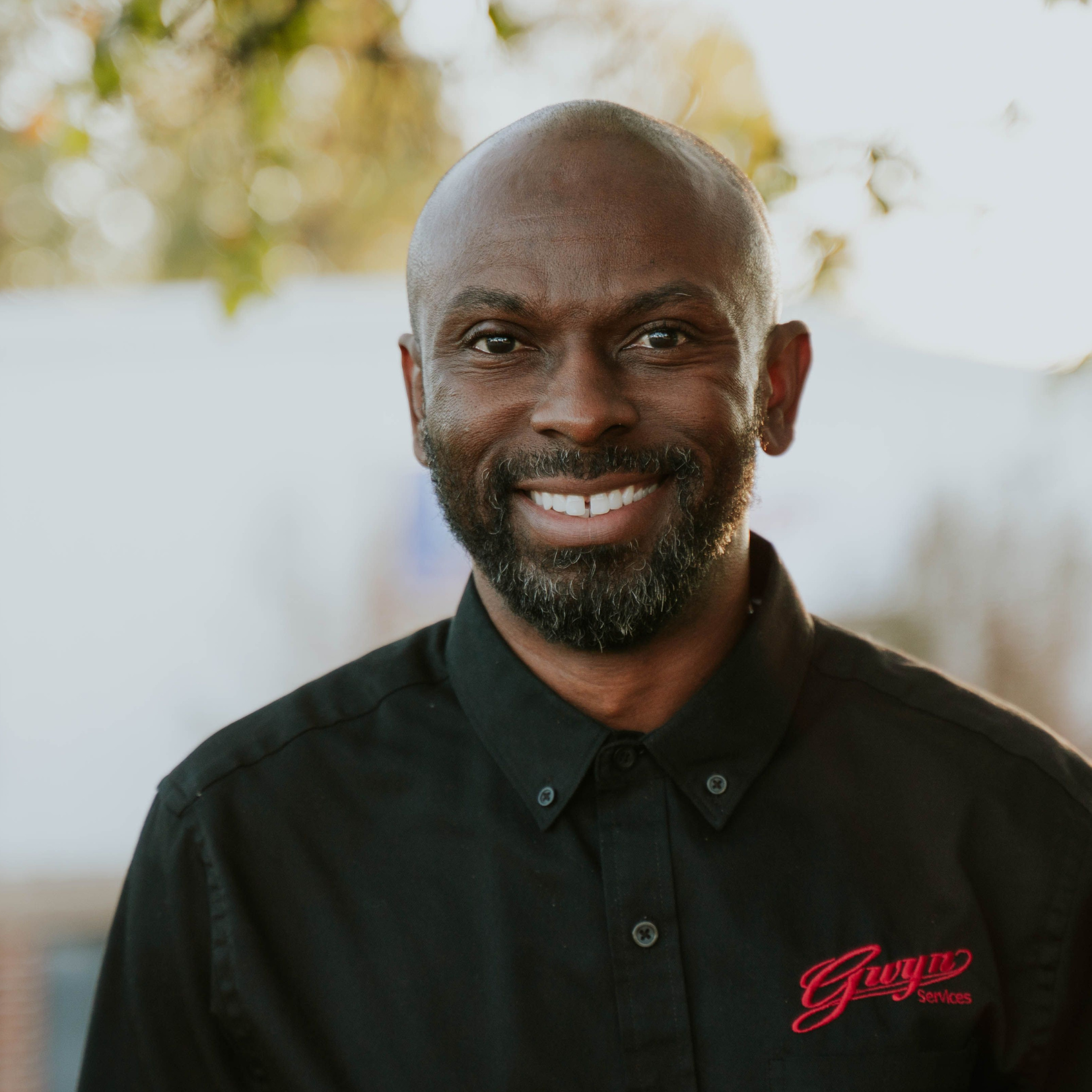 Smiling man in a black shirt with a red logo, standing outdoors against a softly blurred background