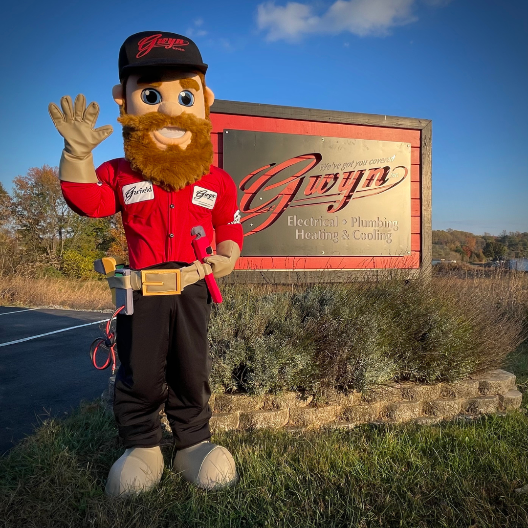 Costumed mascot waving beside a vintage sign in a grassy outdoor field under a blue sky