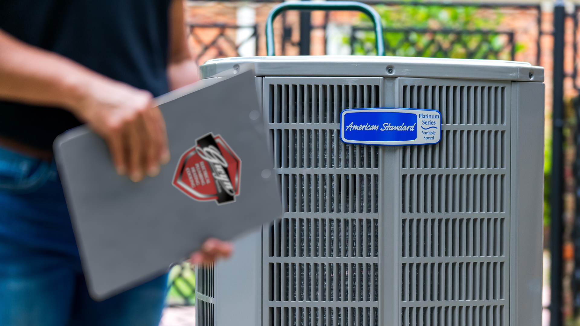 Technician installing or inspecting a gray HVAC unit outdoors, holding a panel with a red warning label.