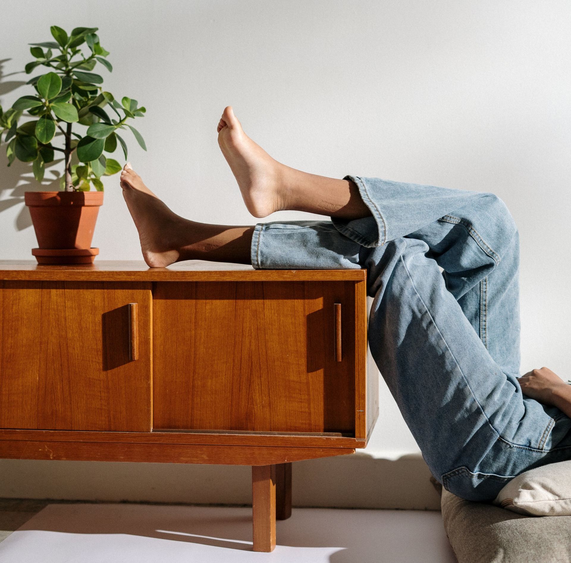 Legs in jeans draped over a wooden cabinet beside a potted plant