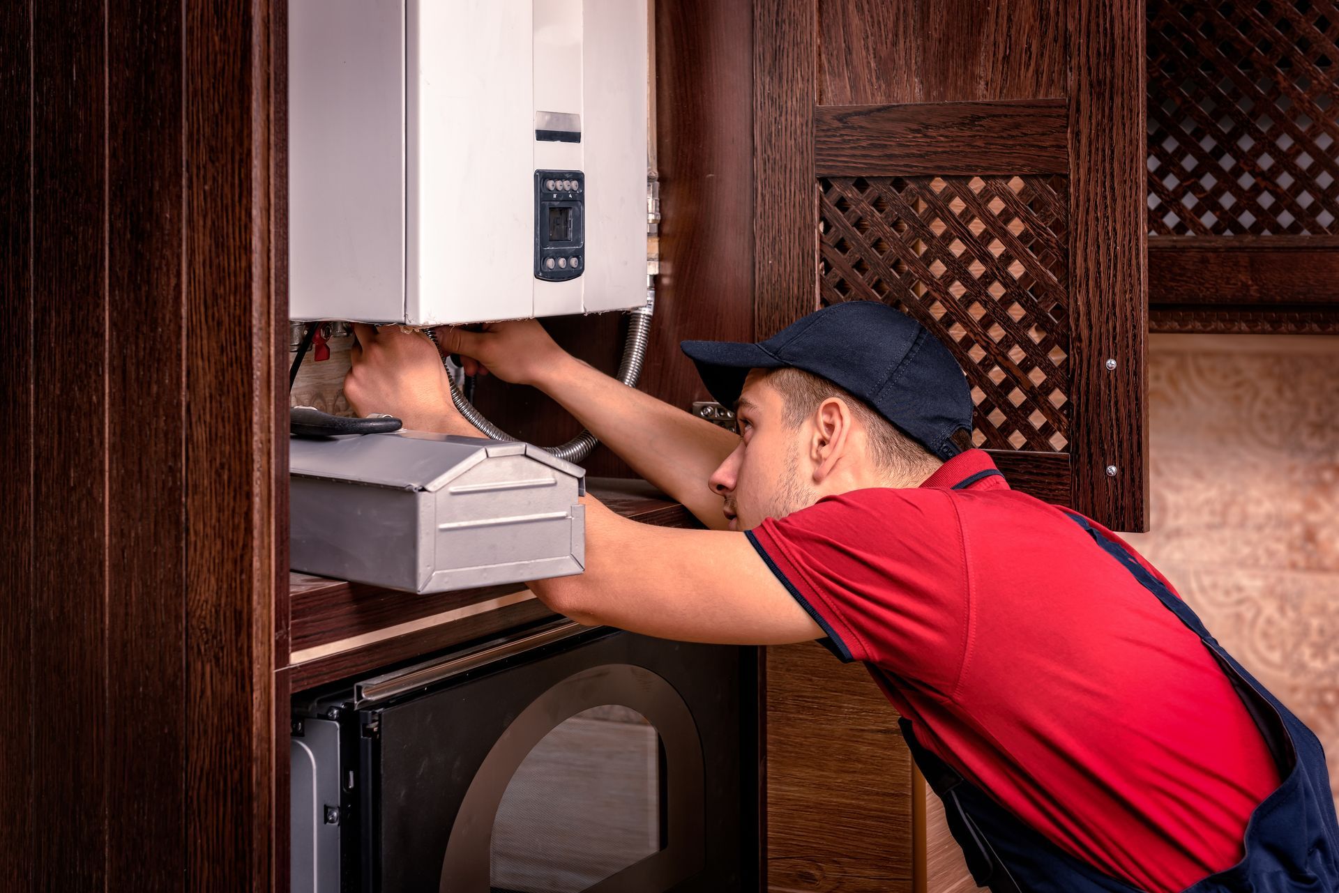 Man in red shirt reaching into a cabinet beneath a wall-mounted appliance in a wood-paneled room