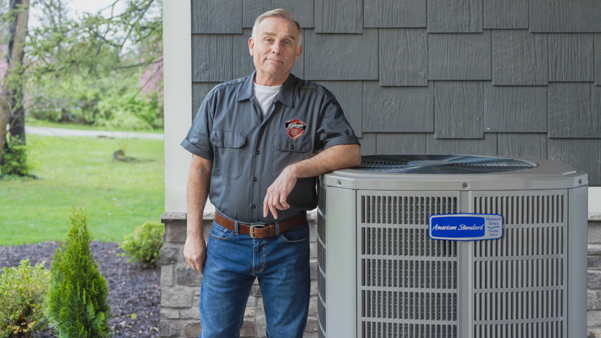 Man standing beside an outdoor HVAC unit on a home patio, with a lawn and shrubs in the background